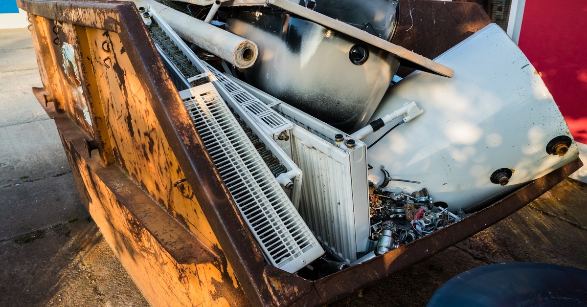 A large yellow dumpster filled to the top with various metal components, including heaters, pipes, and vents.