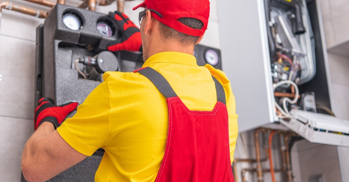A man in a yellow shirt with red overalls and gloves performing a hands-on check on a water heating system.