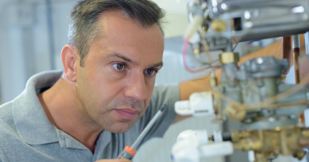 A man in a grey polo shirt holding a screwdriver and looking closely at the inner electrical workings of an appliance.