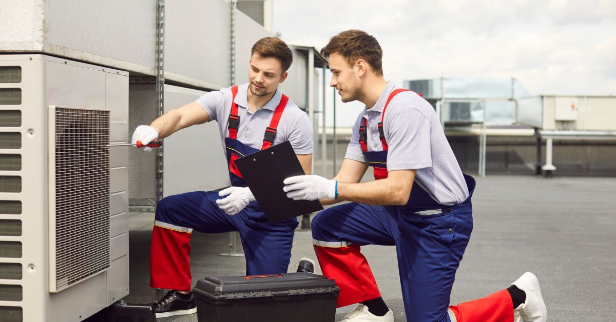 Two men in red and blue overalls kneeling next to an air conditioning unit on a building’s roof. One holds a screwdriver while the other hold a clipboard.