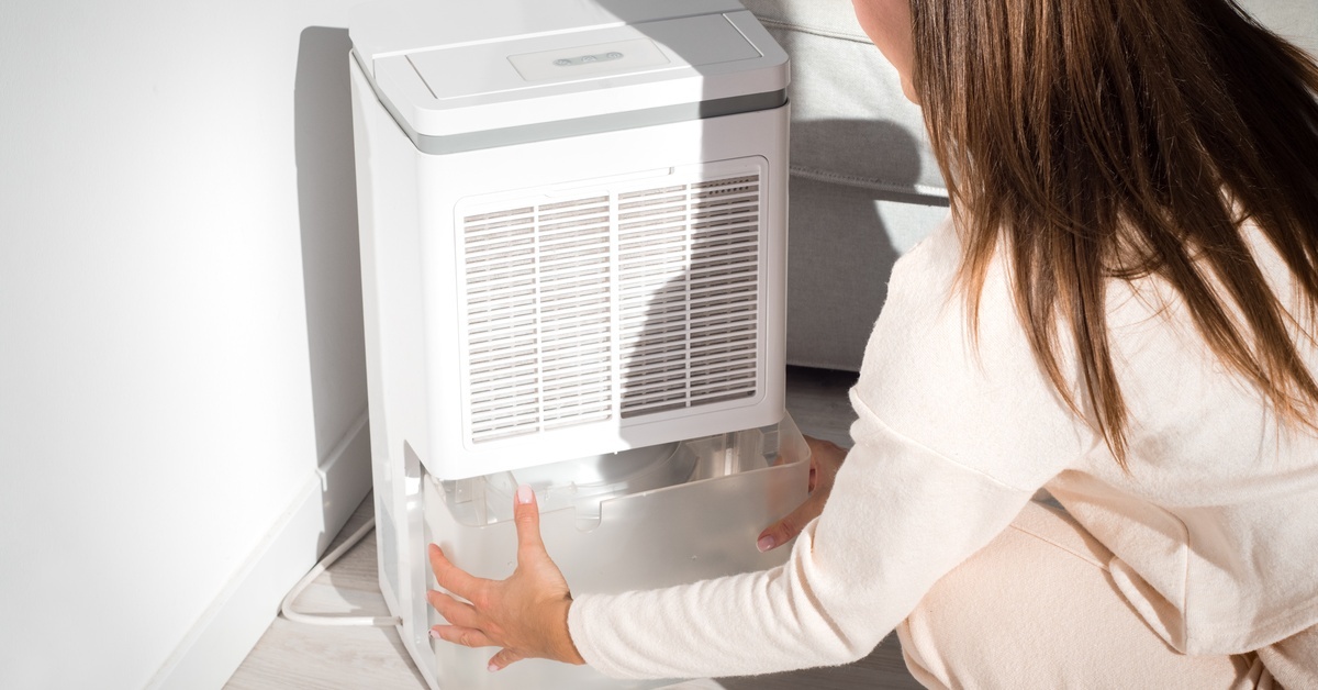 A woman is kneeling down beside a dehumidifier. She’s taking out the tank, which contains water.