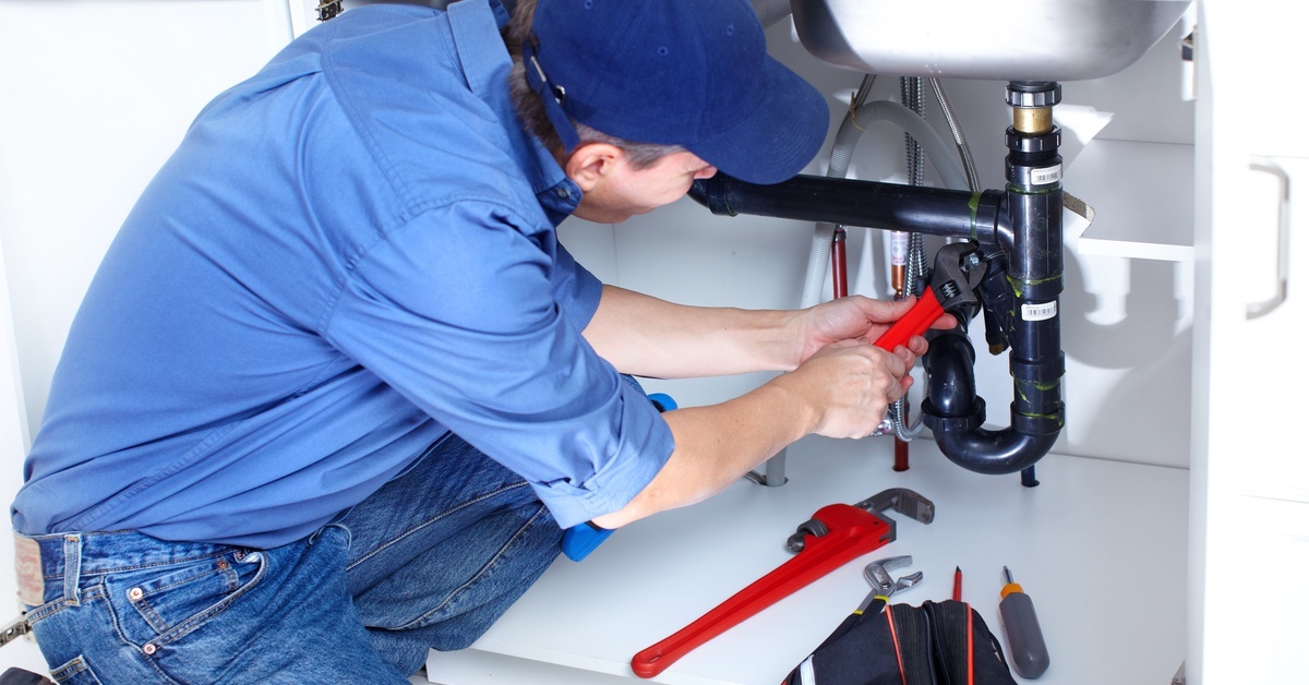 A man in a collared blue shirt, a blue cap, and jeans tightening a nut on a pipe with a wrench. The pipe is black.