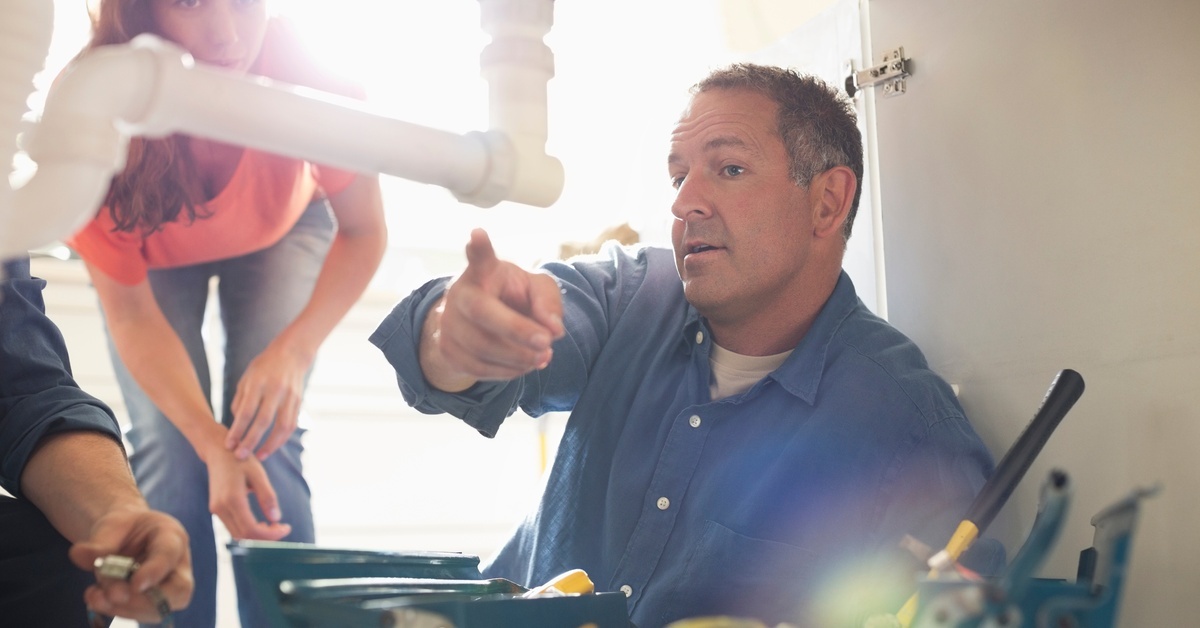 A man in a blue shirt pointing to pipes in a cabinet. A woman standing behind him also looks at the pipes.