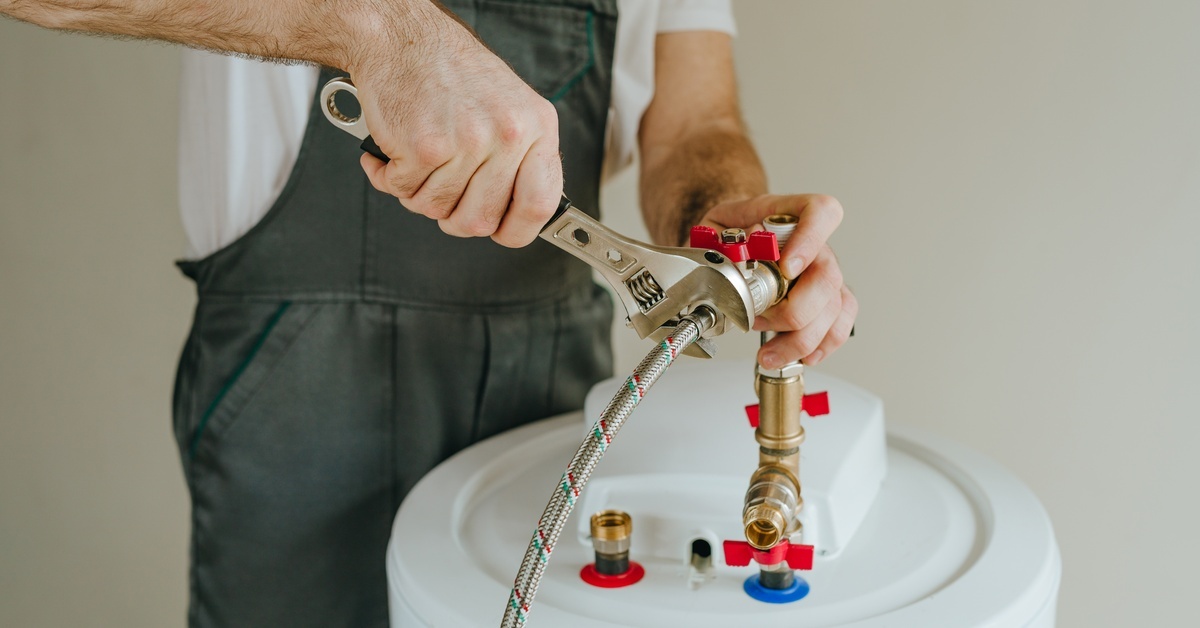 A worker in dark coveralls uses a wrench to tinker with the piping of a classic tank-style water heater.