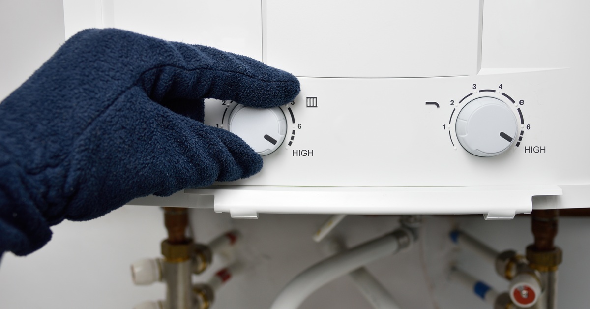 A white water heater mounted on a wall. A person’s gloved hand is turning a knob on the water heater.
