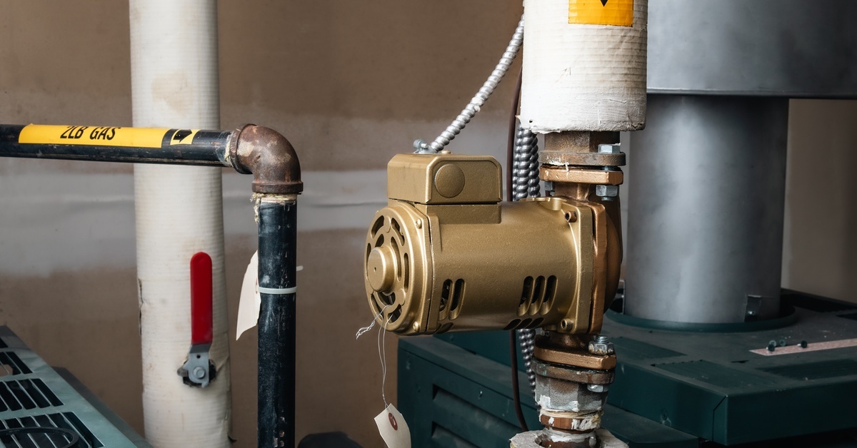 A small, gold-colored booster pump attached to a system of water pipes. There is a hot water tank and gas lines nearby.
