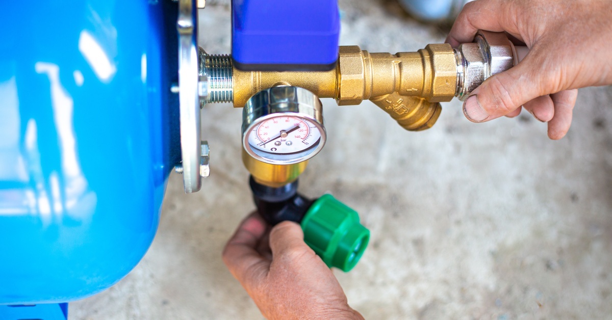 A person’s hands holding onto two connections attached to a large blue pump. A pressure gauge is visible near the top of the connections.