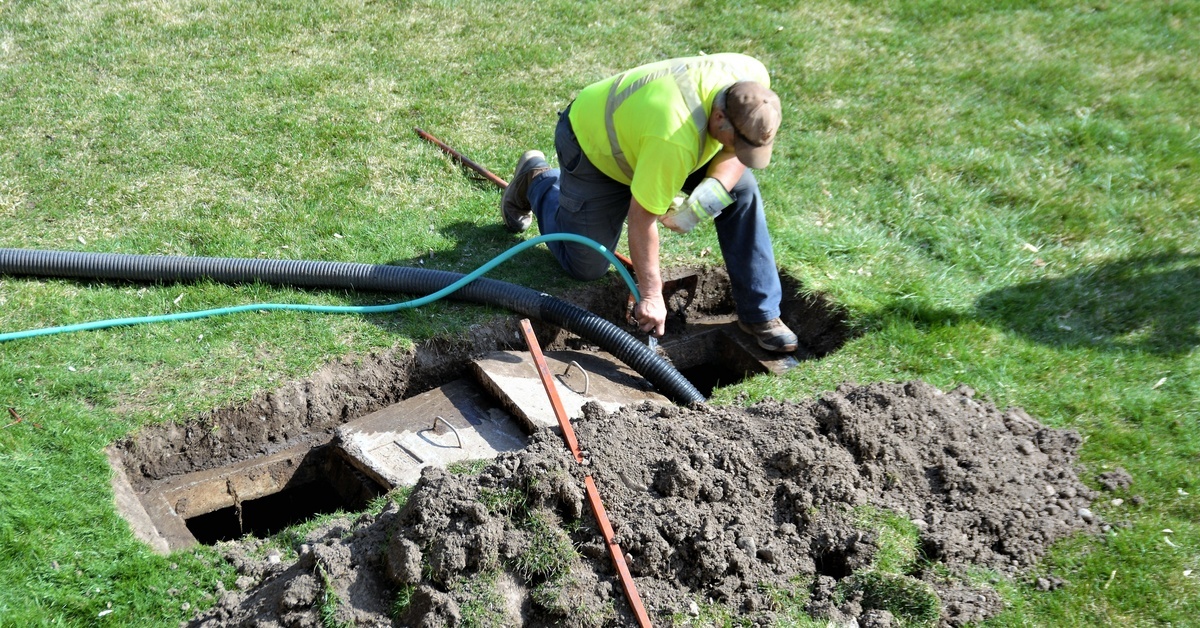 A man in a hi-vis vest is kneeling over a dug hole in the ground. There is a pipe coming out of the ground.