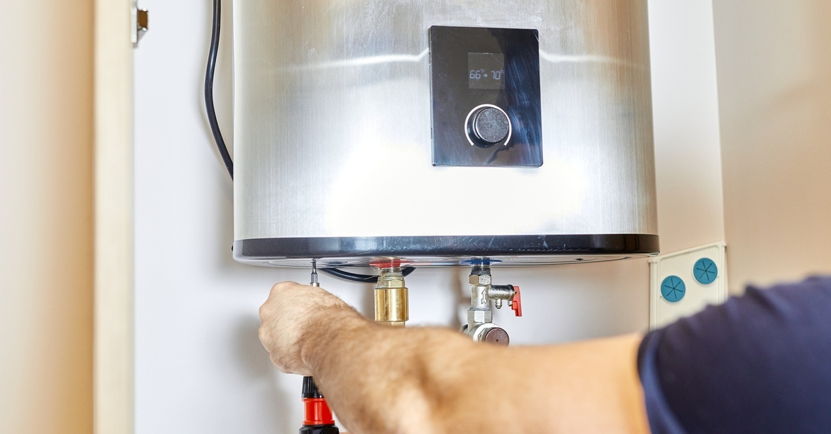 A person reaches forward with a screwdriver in their hand to adjust part of a sleek silver tankless heater.