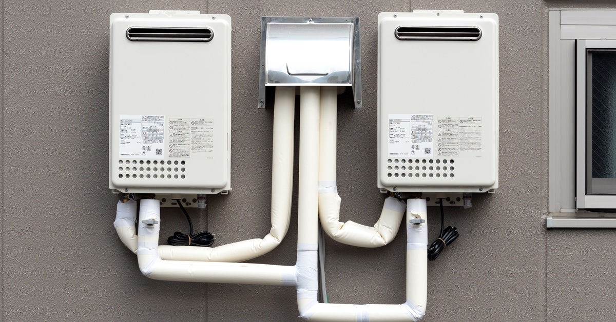 Two small tankless water heaters are mounted on a wall, with white flexible piping connecting the two units.