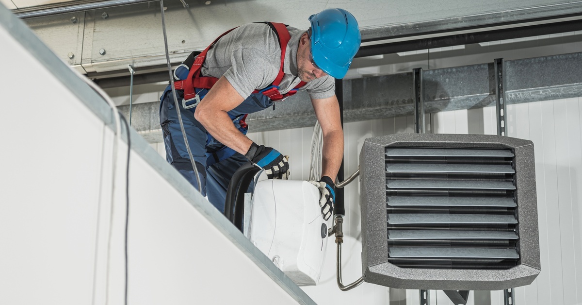A man in denim overalls and a blue hard hat stands on a high platform to install a new water heater.