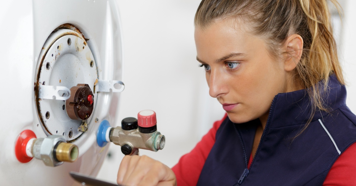 A woman in a red shirt and blue vest holds a digital tablet as she examines the workings of a hot water heater.