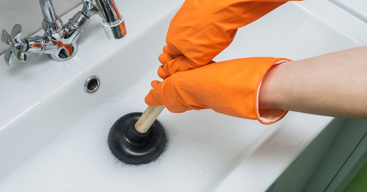 A person wearing orange gloves using a small handheld plunger to unclog the drain of a rectangular sink.