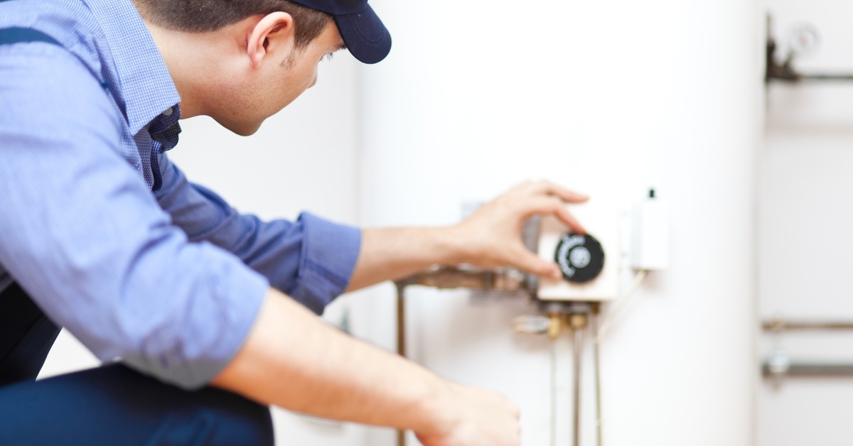 A plumber in a blue hat, shirt, and pants kneeling down while checking the dial on the side of a water heater tank.